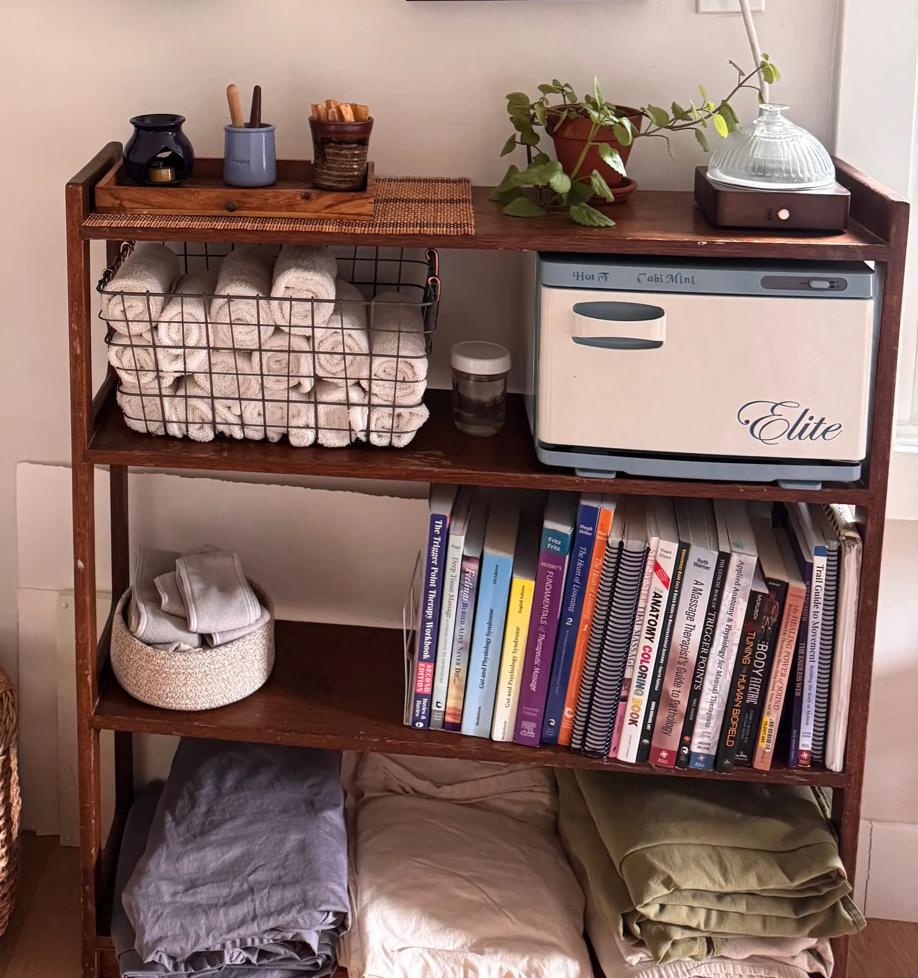 A shelf of warm rolled towels, a towel warmer, folded linens, and Briana's anatomy and bodywork texts
