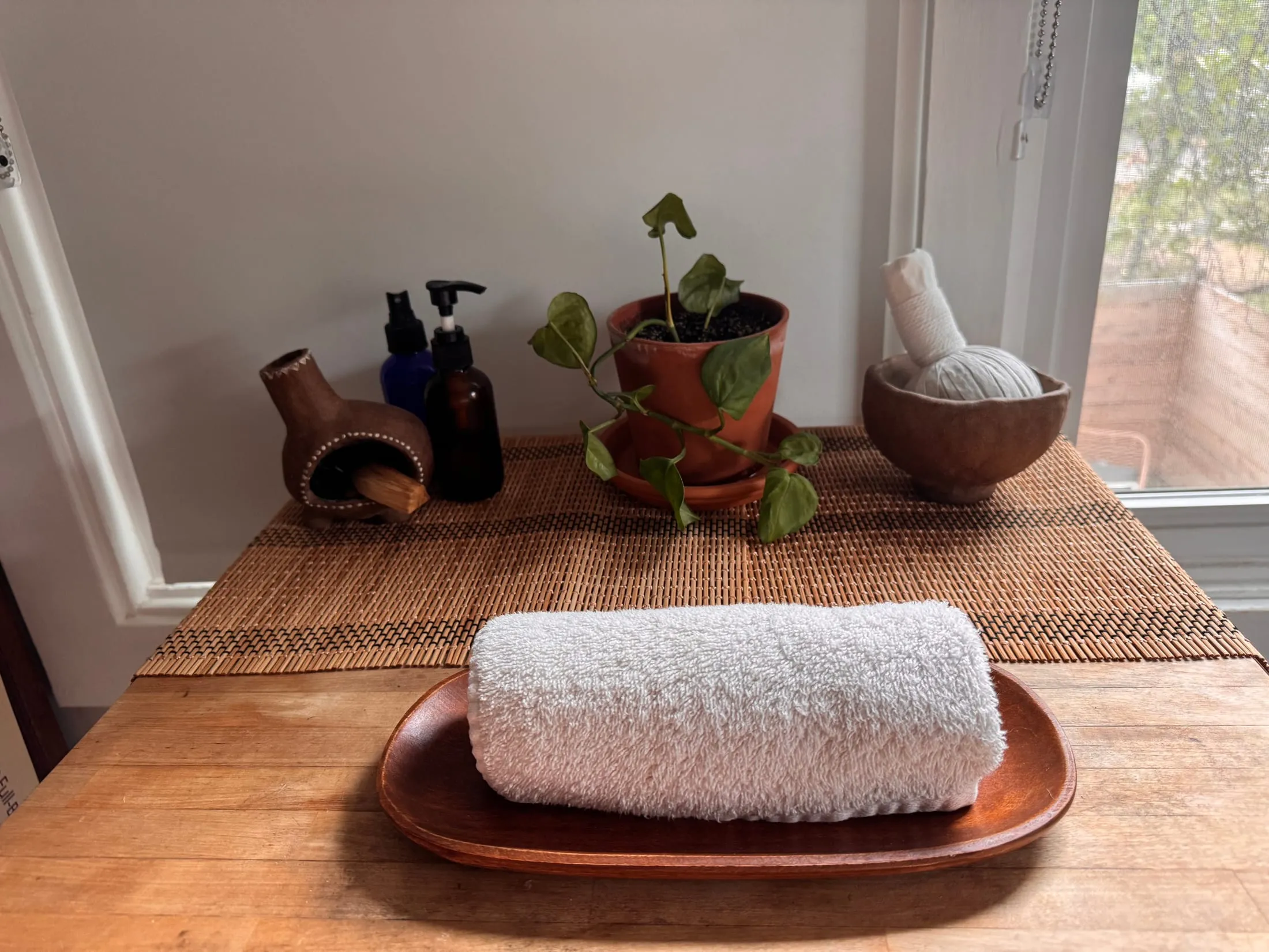 A rolled towel on a wooden tray, with massage oils, a herbal compress, and a trailing pothos